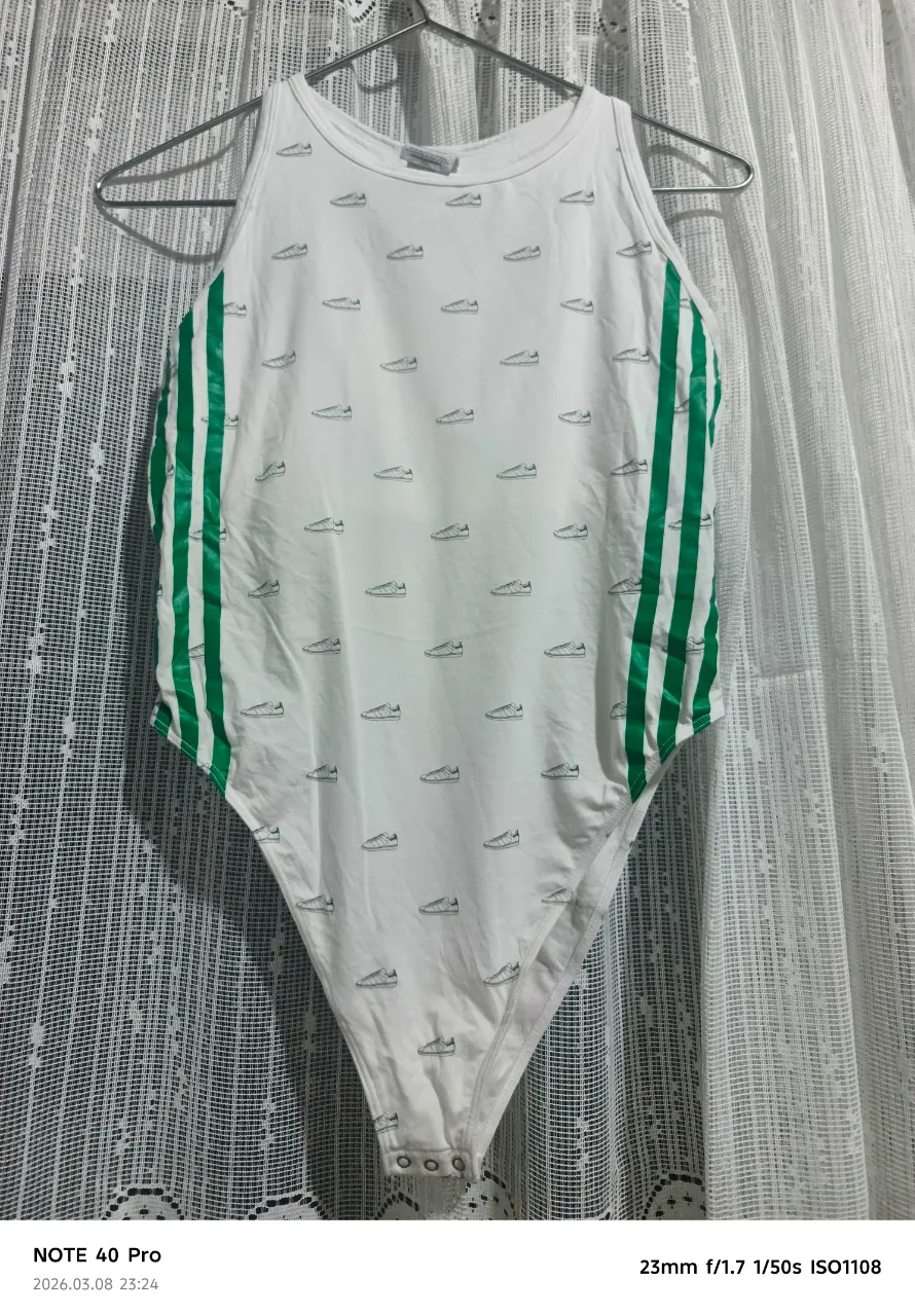 Body de algodón blanco con estampa de zapatillas Adidas en verde. Tiene las tres tiras verdes icónicas de la marca en los laterales. Ideal para un look deportivo y canchero.