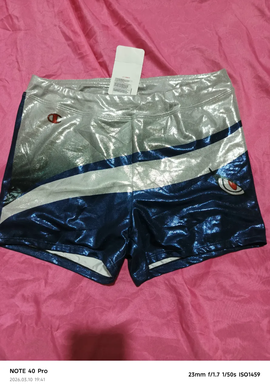 Shorts deportivos con diseño de franjas plateadas y azules brillantes. Presenta un logo de Champion en la parte superior izquierda y un diseño de ojo estilizado en la parte inferior derecha.