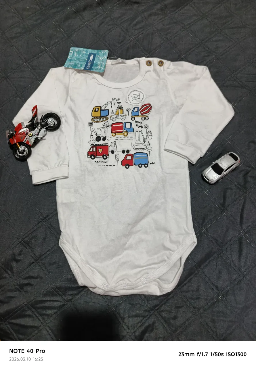 Body de manga larga blanco con estampa de vehículos de construcción y camiones en la parte delantera. Tiene broches en la parte inferior para facilitar el cambio de pañal.