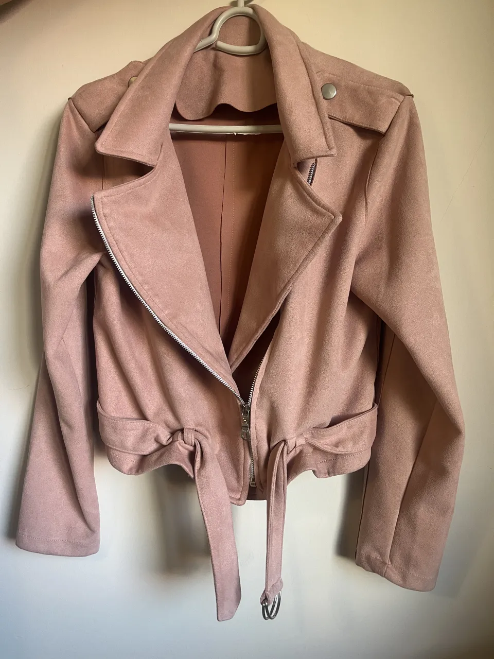 Campera de gamuza rosa, estilo biker, con cinturón para anudar a la cintura. Posee cierre metálico al frente y solapas. 