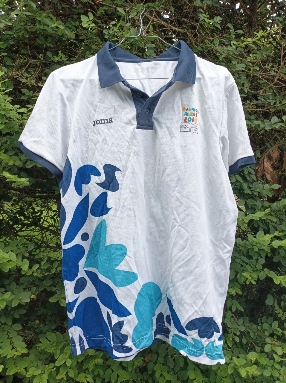 Remera deportiva blanca con estampa de hojas en tonos azules y celestes. Cuello polo con botones y detalles en azul marino. Ideal para entrenamientos o uso casual.