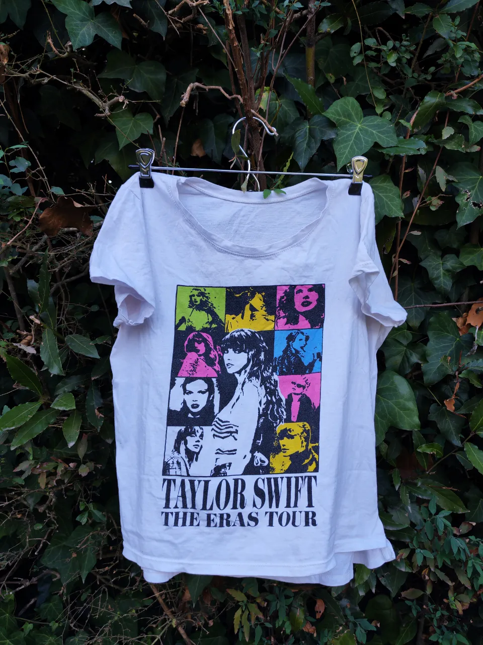 Remera blanca de algodón con estampa "Taylor Swift The Eras Tour". La estampa es un collage de fotos de la artista en diferentes colores y estilos. Ideal para fans de Taylor Swift y para usar en recitales o eventos.