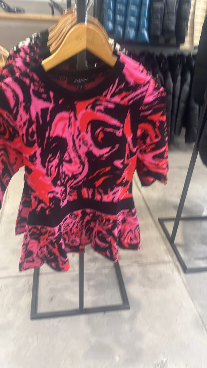 Sweater estampado con diseño abstracto en fucsia, rojo y negro. Posee cuello redondo y mangas cortas. La parte inferior tiene un corte con volado.