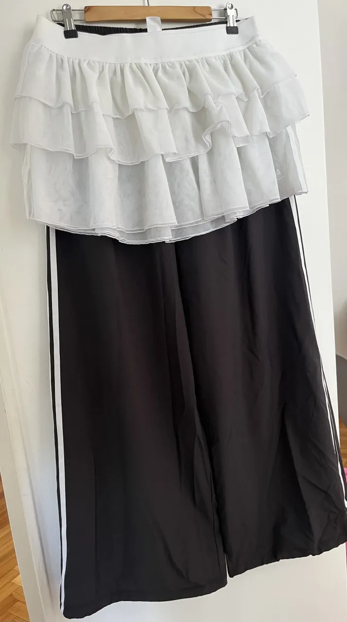 Conjunto de pollera y pantalón de Adidas. La pollera es blanca, con volados y detalles de la marca. El pantalón es negro, ancho, con las tres tiras clásicas de Adidas a los costados. Ideal para un look deportivo y canchero.