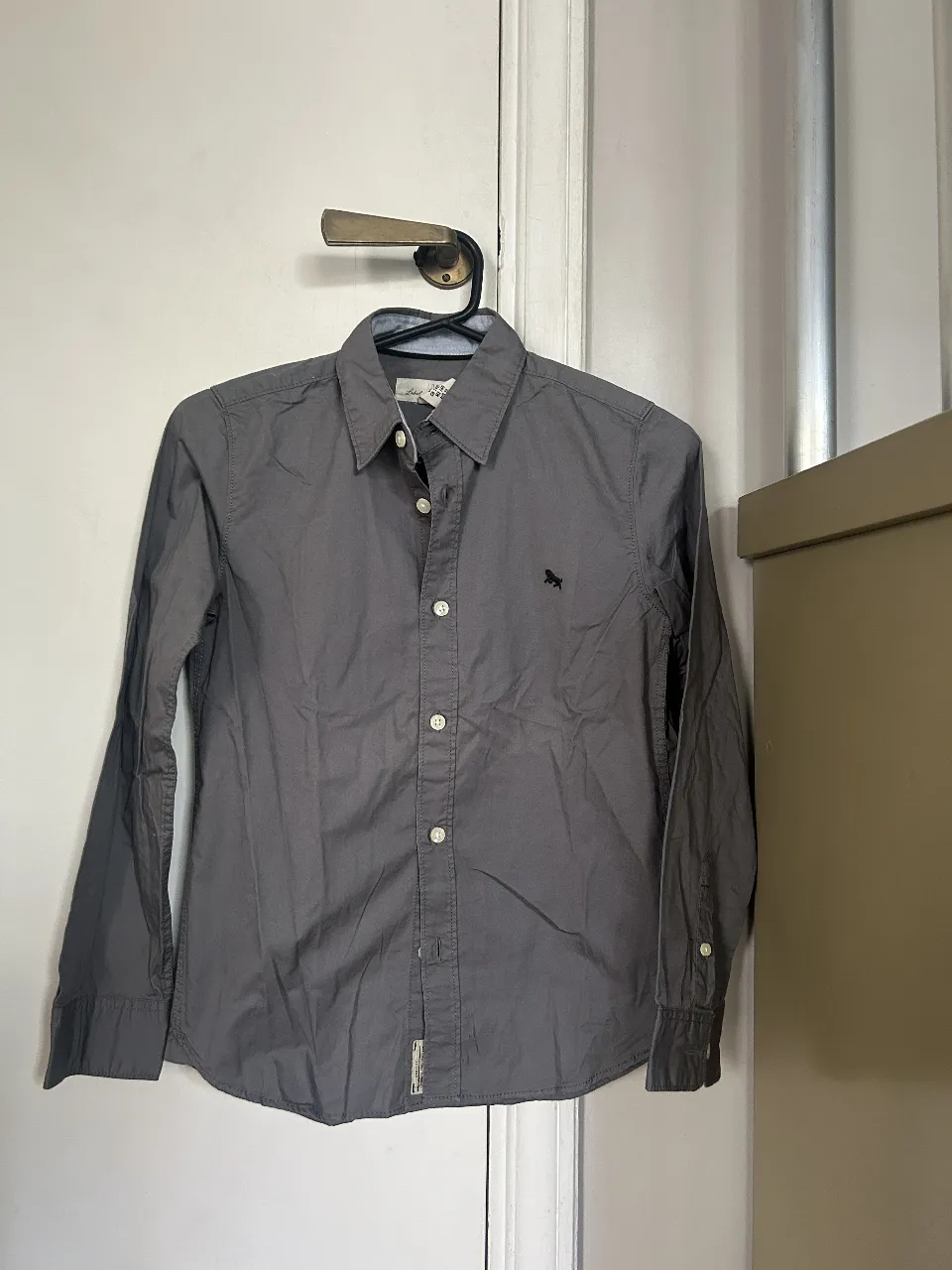 Camisa gris de niño, manga larga, con logo bordado en el pecho. Ideal para un look casual y versátil.