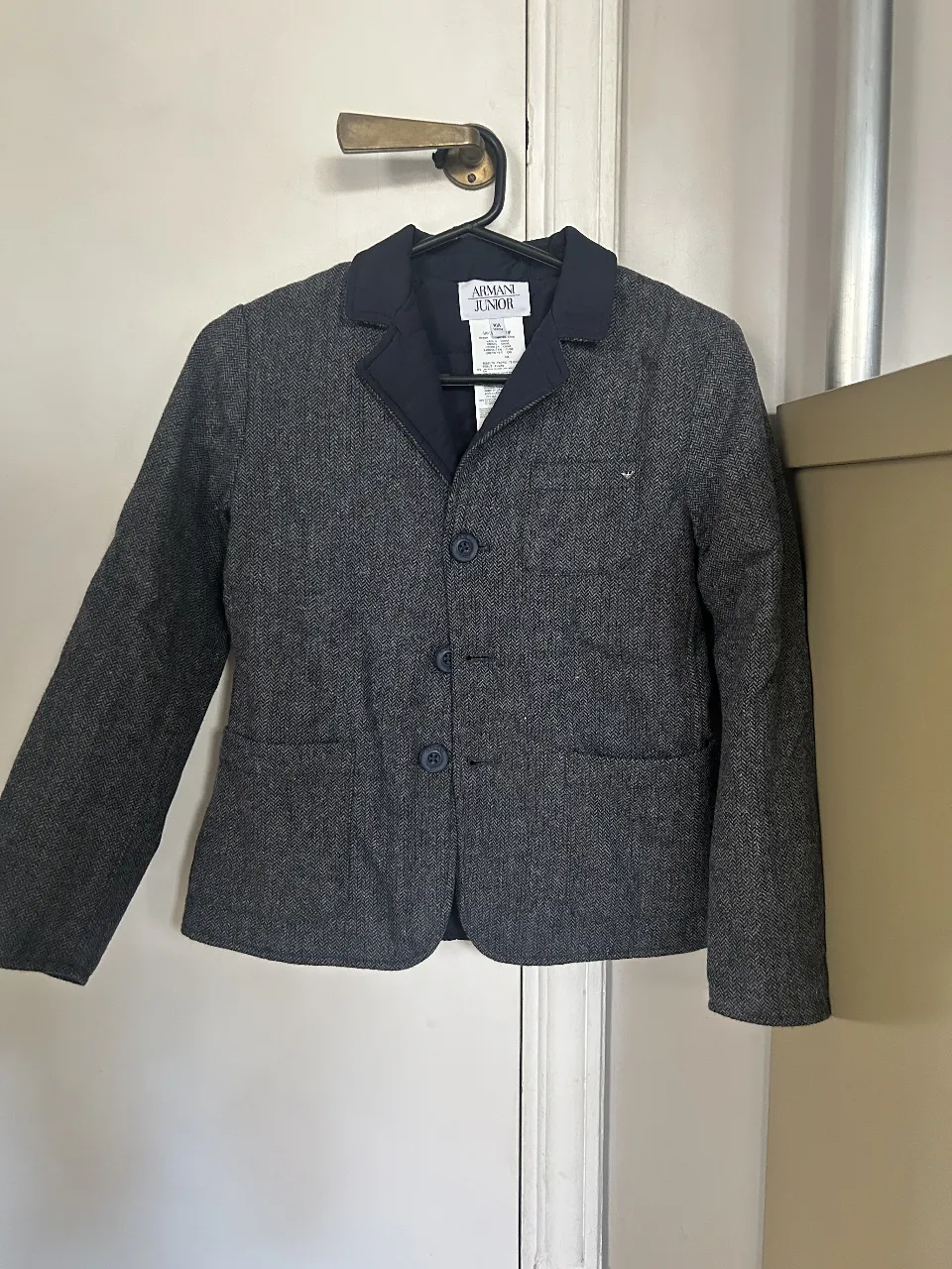 Chaqueta blazer Armani Junior para niño, de color gris con detalles en azul marino. Confeccionada en tejido de lana y viscosa, ideal para ocasiones formales o semi-formales. Cuenta con dos bolsillos frontales y cierre con botones.