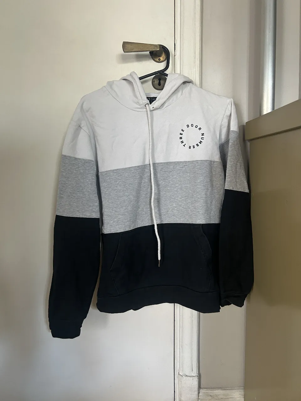 Buzo canguro tricolor para hombre, con capucha y bolsillo frontal. Diseño en blanco, gris y negro. Ideal para un look casual y cómodo.