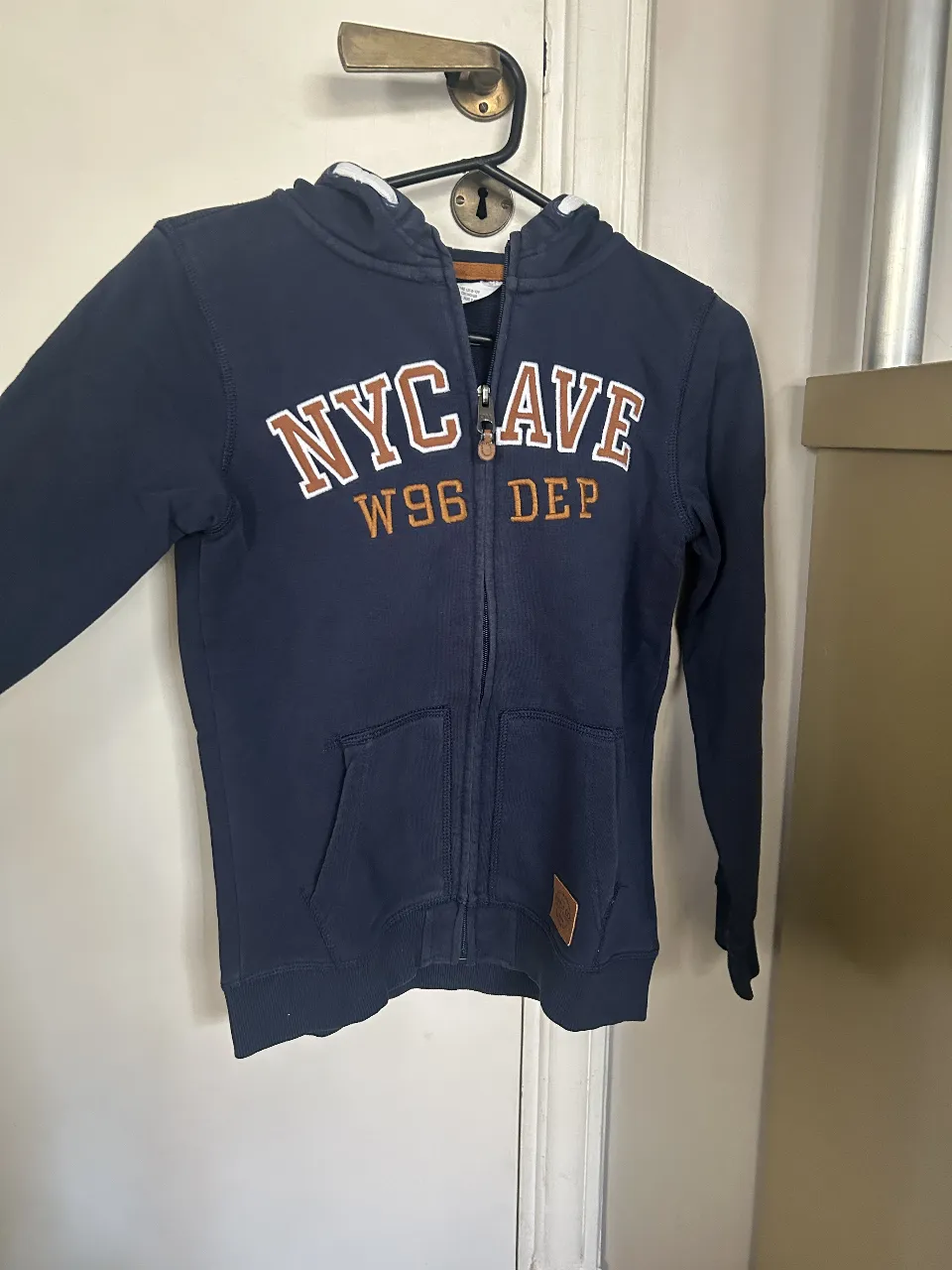 Buzo con cierre de la marca H&M, de color azul marino con detalles en marrón. Tiene capucha y el estampado "NYC AVE W96 DEP" en el frente. Ideal para un look casual y abrigado.