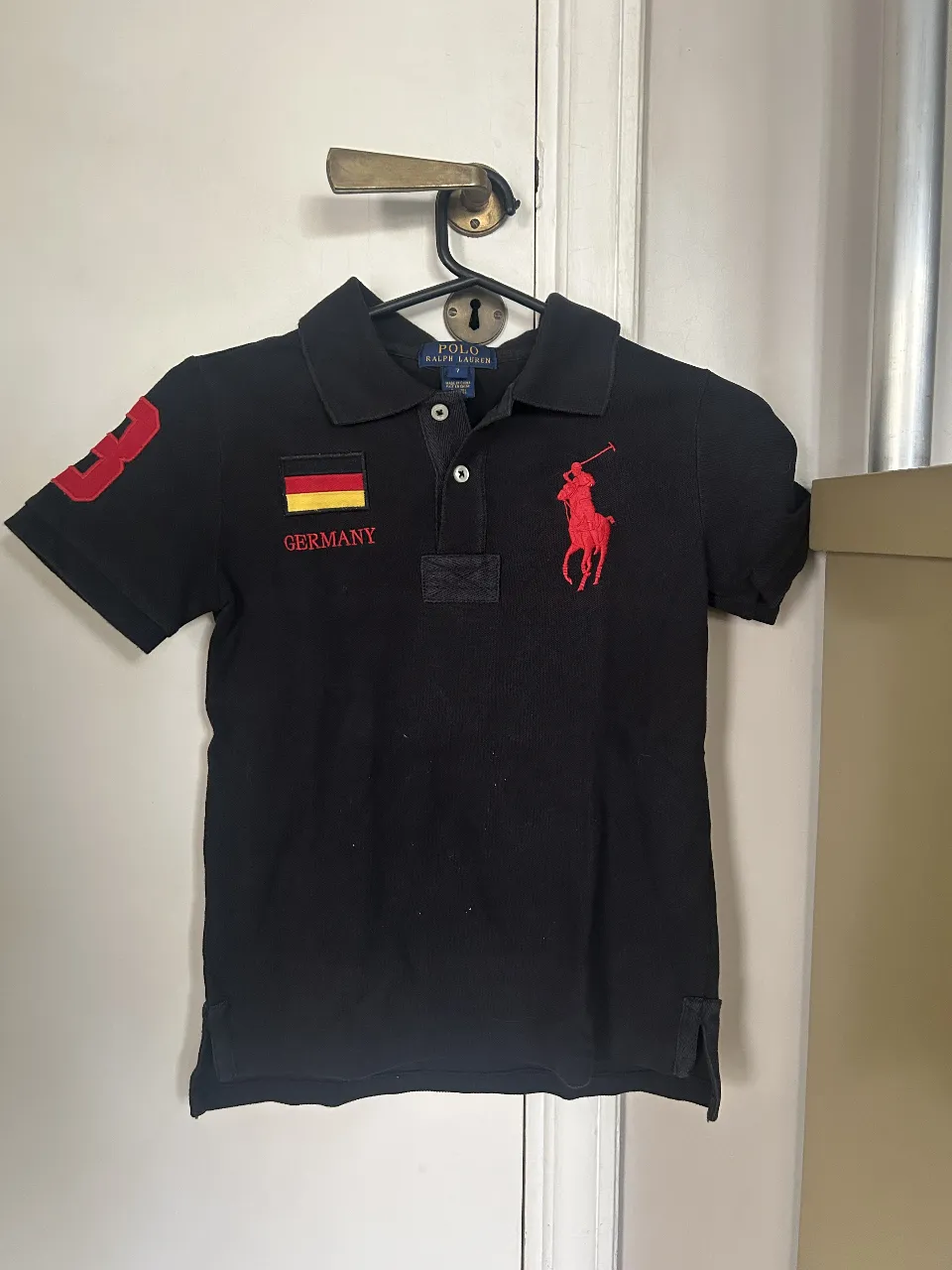 Remera polo negra de niño, con el número 3 y la bandera de Alemania en rojo y amarillo. Logo del caballo rojo de Ralph Lauren en el pecho. Ideal para un look casual y deportivo.