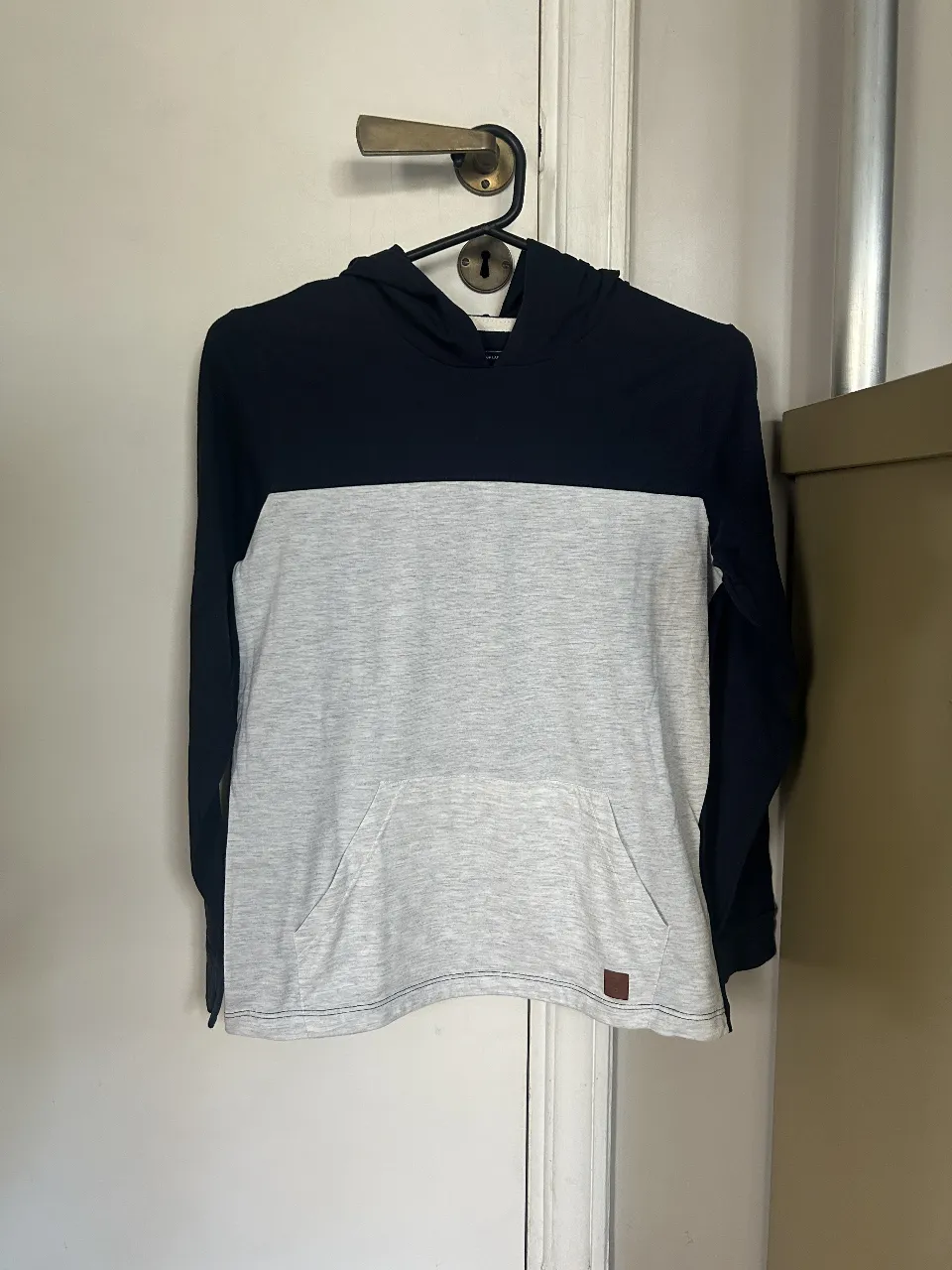 Buzo con capucha para hombre, de color azul marino y gris claro. Confeccionado en tela suave y cómoda, ideal para uso diario. Posee bolsillo tipo canguro y el logo de la marca en la parte inferior.