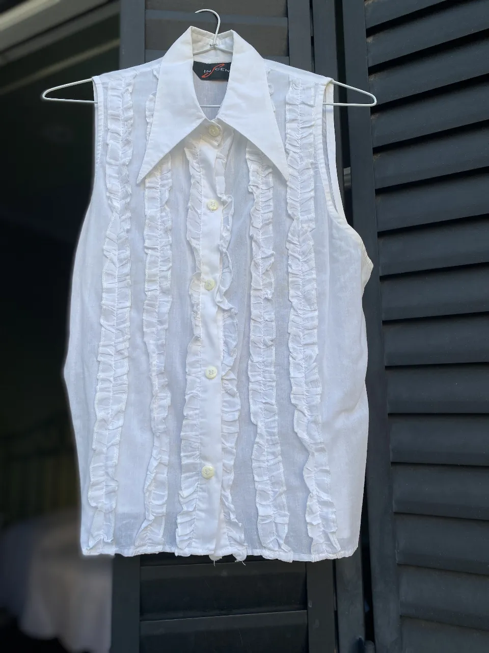 Blusa blanca sin mangas con detalles de volantes verticales. Cuello camisero y cierre con botones. Ideal para un look elegante y fresco.
