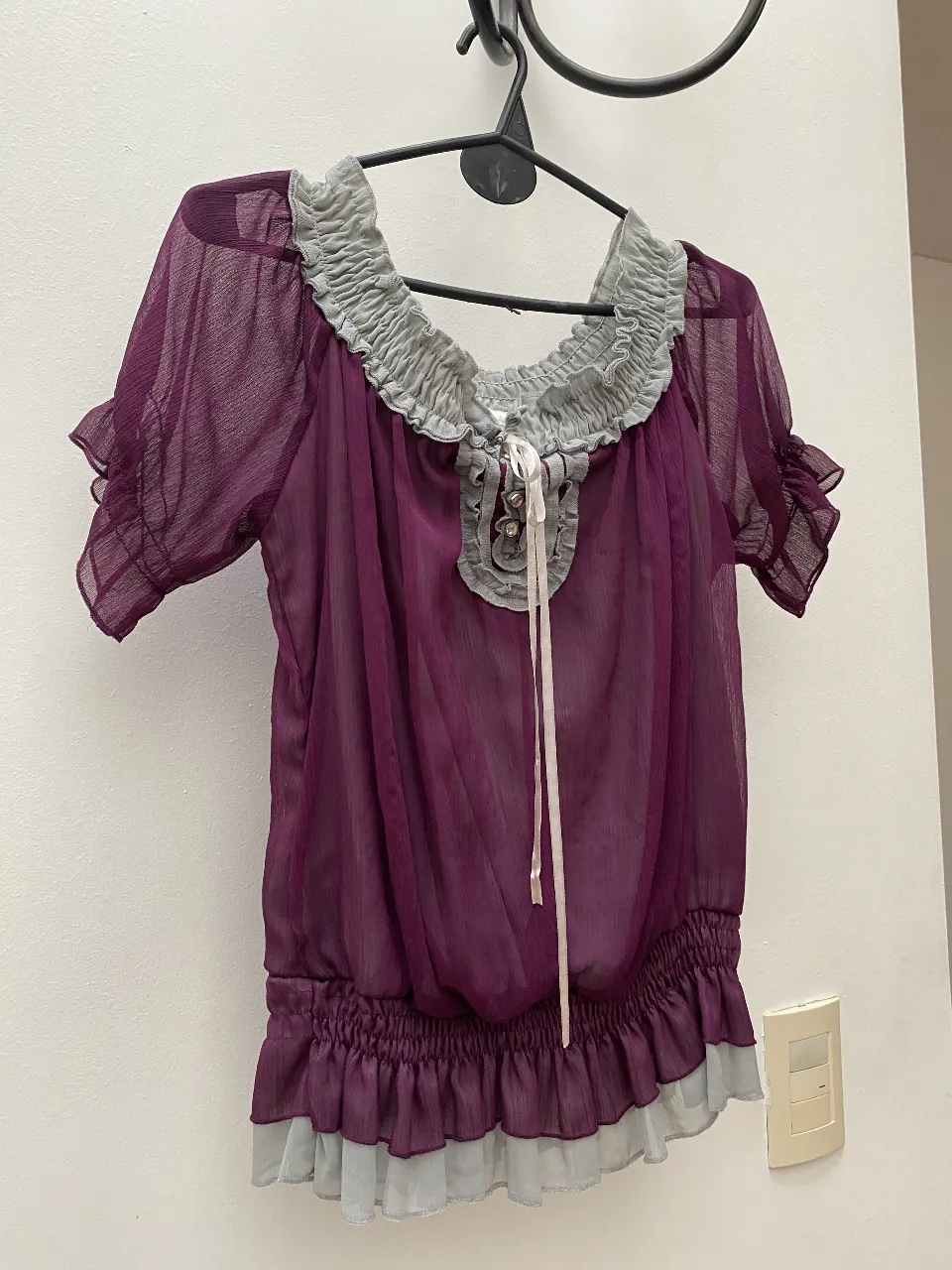 Blusa de gasa color morado con detalles grises en el cuello y el bajo. Tiene mangas cortas con volados y un lazo en el frente. Ideal para un look elegante y femenino.