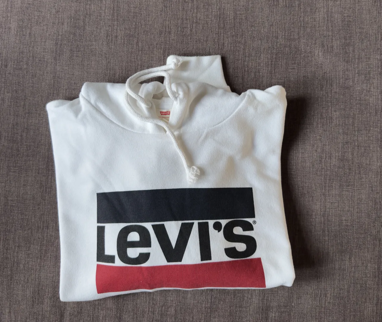 Buzo blanco con capucha y cordones. Estampa frontal con el logo de Levi's en negro, blanco y rojo.
El logo también se encuentra  en una de las mangas.