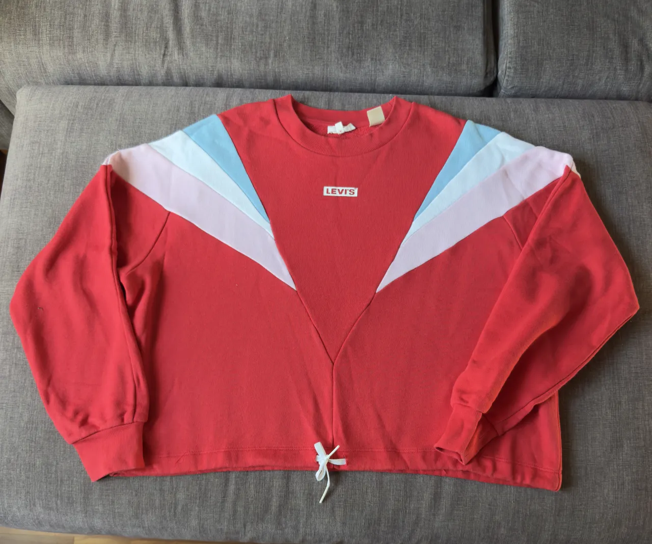 Buzo crop de algodón color rojo con detalles de bloques de color en los hombros en blanco, rosa pálido y celeste. Posee un cordón ajustable en la cintura. Logo de la marca en el frente.