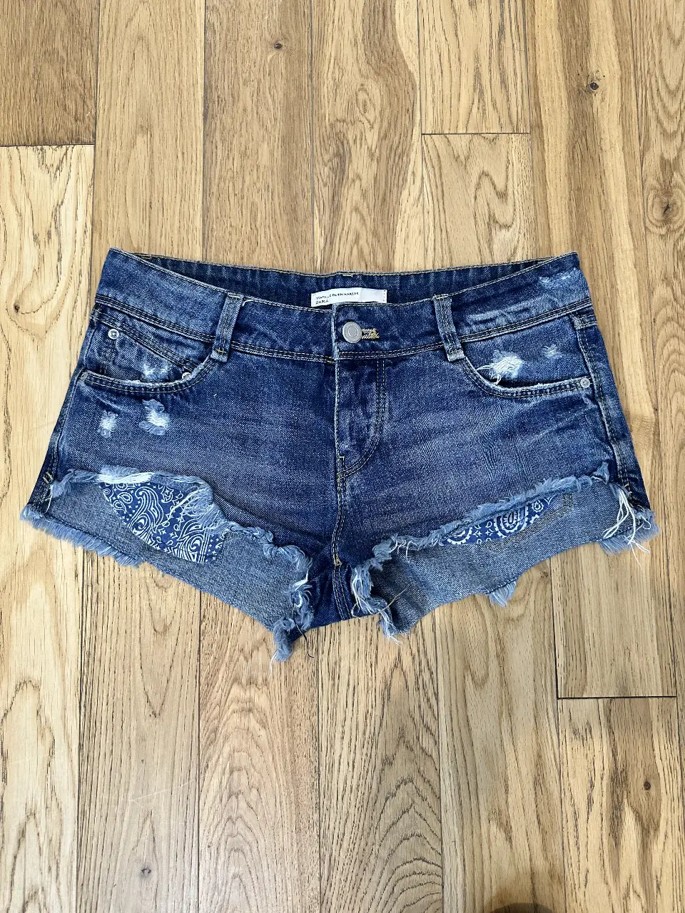 Short de jean azul ZARA con detalles rotos y bordados en los laterales. Tiro medio y corte desflecado. Ideal para un look casual y moderno.