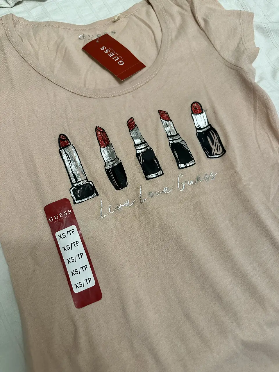 Remera Guess de mujer, color rosa claro, con estampado de labiales con brillos y la frase "Live Love Guess". Ideal para un look casual y moderno.