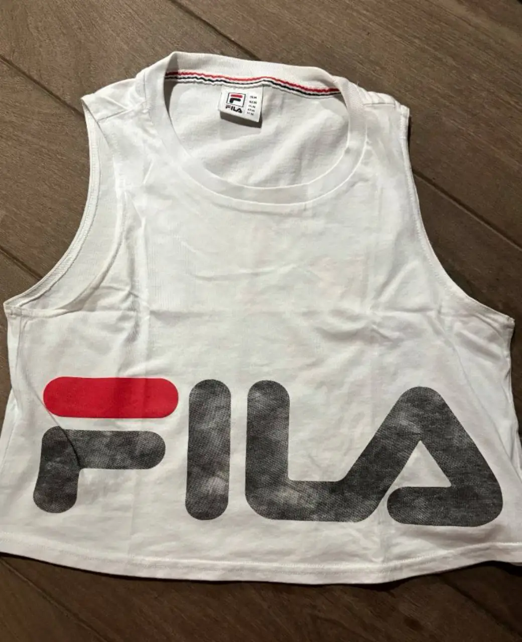 Musculosa deportiva blanca de la marca Fila, con el logo estampado en el frente. Ideal para entrenar o para un look casual y cómodo. Es estilo pupera, corta 