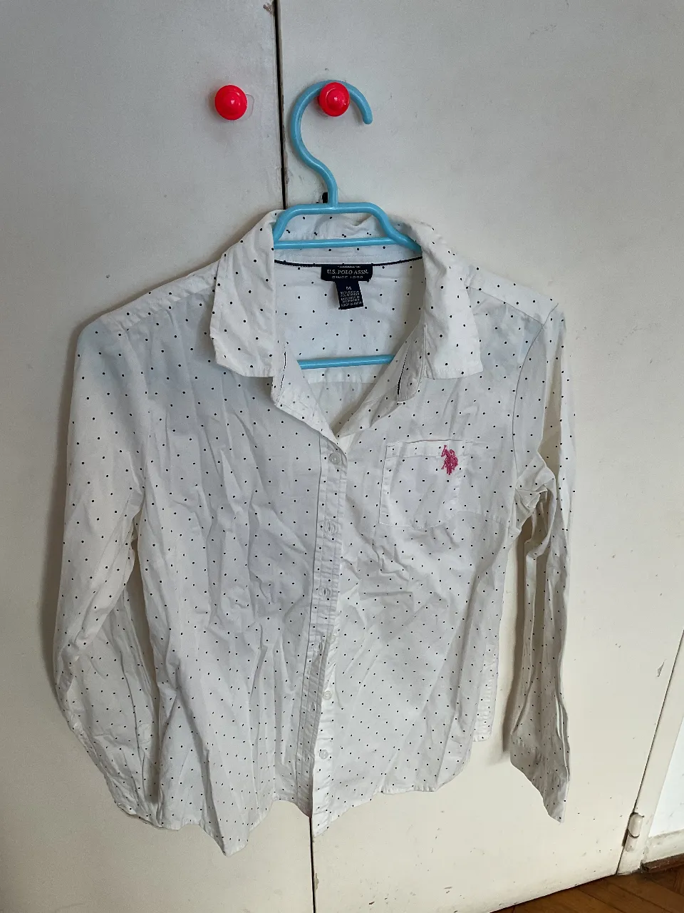 Camisa de mujer blanca con estampado de lunares negros, de la marca U.S. Polo Assn. Confeccionada en algodón y spandex, manga larga y un bolsillo en el pecho. Ideal para un look casual y versátil.