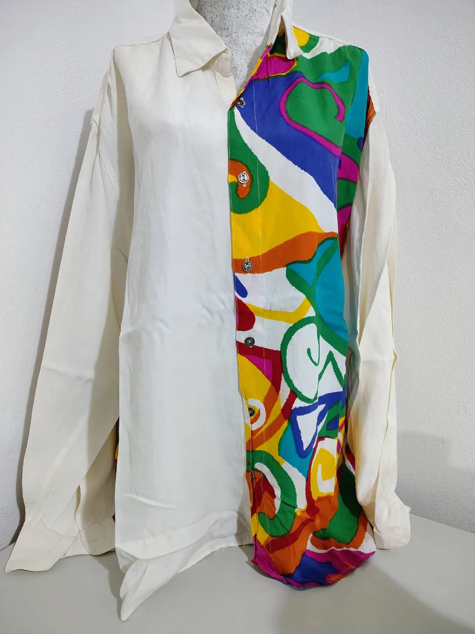 Camisa oversize de manga larga, con un diseño único y llamativo. La mitad de la prenda es lisa en color blanco roto, mientras que la otra mitad presenta un estampado abstracto multicolor vibrante. Ideal para un look audaz y original.