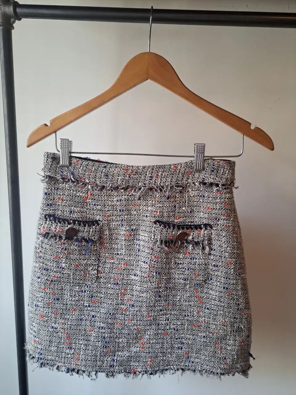 Pollera de tela tweed con detalles en azul y naranja. Diseño con bolsillos delanteros y botones decorativos. Ideal para un look elegante y sofisticado.