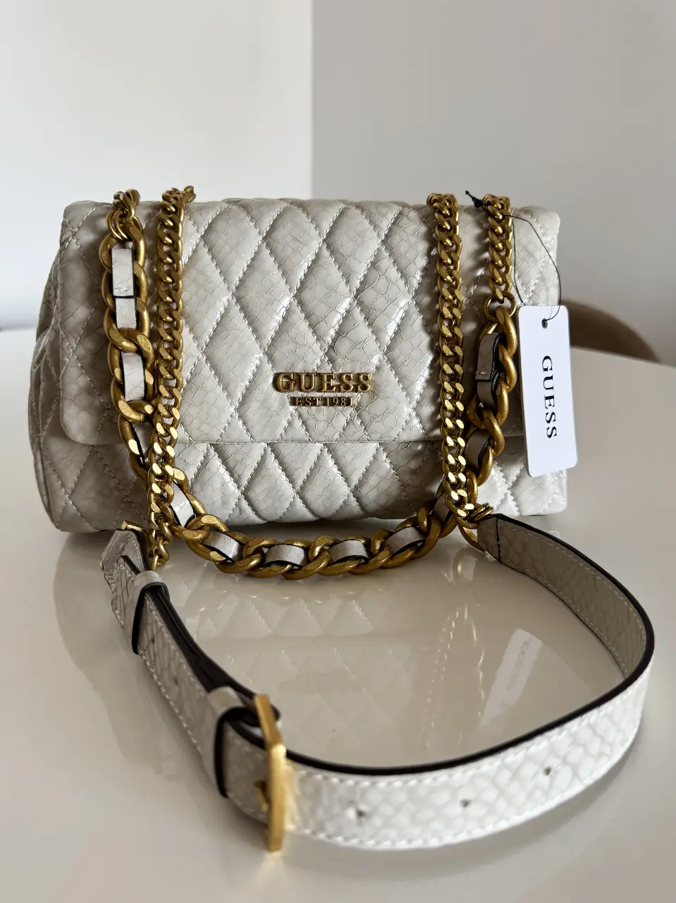 Cartera Guess Importada color crudo con detalles y cadena en color dorado. Diseño acolchado con textura símil piel de serpiente. Ideal para un look elegante y moderno.