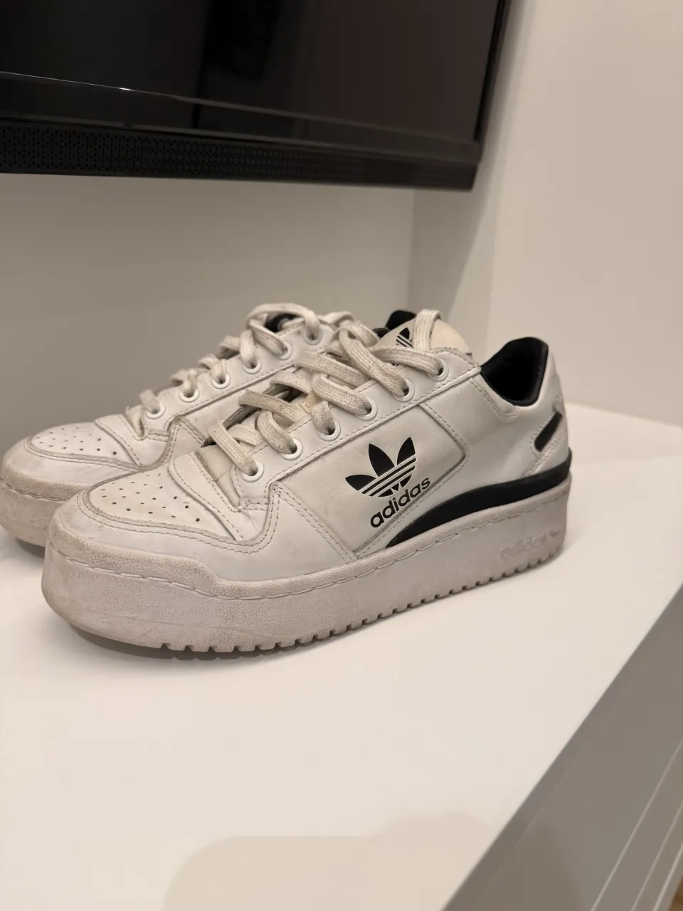Zapatillas Adidas blancas de cuero con detalles en negro y suela gruesa. Tienen cordones blancos y el logo de la marca en los laterales.