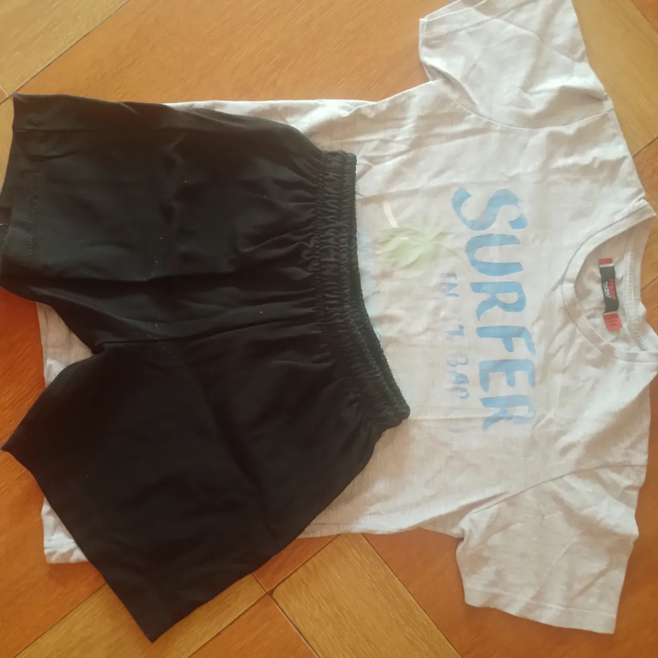 Conjunto de remera y short de algodón, ideal para el verano. La remera es de color gris claro con estampa "SURFER" en azul y verde, y el short es negro liso. Perfecto para un look casual y cómodo.