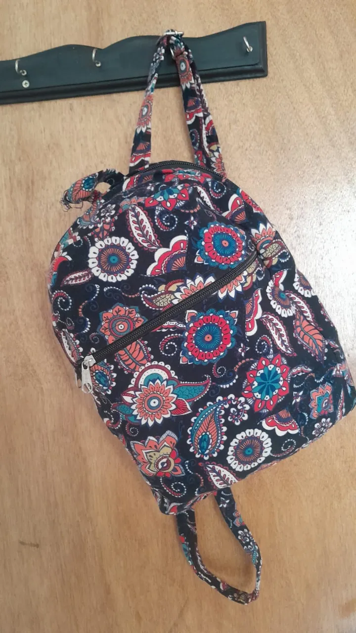 Mochila pequeña con estampado floral y paisleys en tonos azules, rojos, naranjas y blancos sobre fondo negro. Posee dos cierres principales y tiras ajustables.