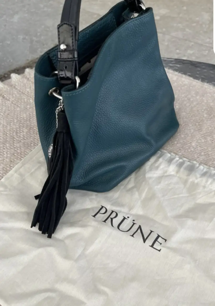 Cartera de hombro PRÜNE de cuero, color verde oscuro. Diseño bucket con detalle de borla negra y correa ajustable. Incluye bolsa guardapolvo.