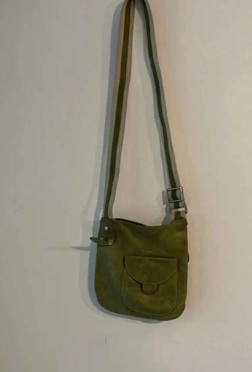 Bandolera de gamuza verde con solapa y bolsillo frontal. Correa ajustable con detalles en beige y hebillas metálicas. Ideal para un look casual y con estilo.