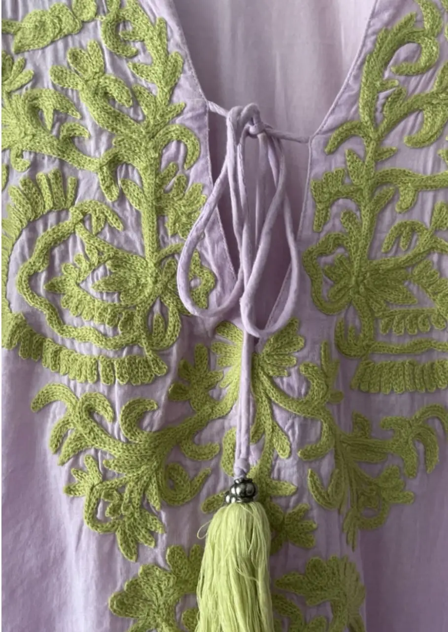 Blusa de mujer estilo boho, con bordados florales en tonos verdes sobre un fondo lila. Tiene mangas 3/4 y detalles de borlas. Ideal para un look casual y fresco.