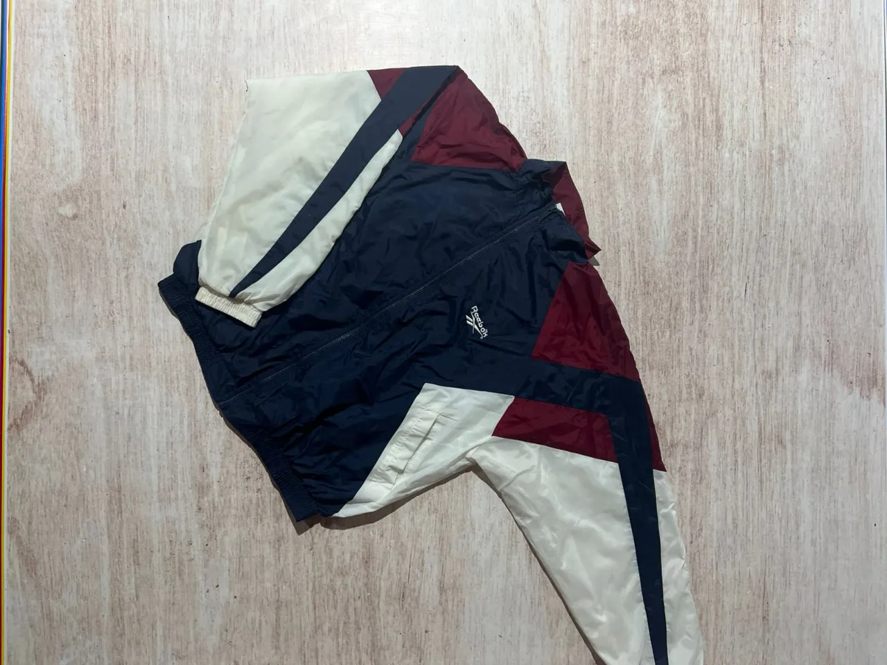 Conjunto deportivo vintage de campera y pantalón, ideal para entrenamientos o un look urbano retro. Diseño con bloques de color en azul marino, granate y blanco roto, con el logo de la marca bordado en la campera. Perfecto para quienes buscan un estilo único y cómodo.