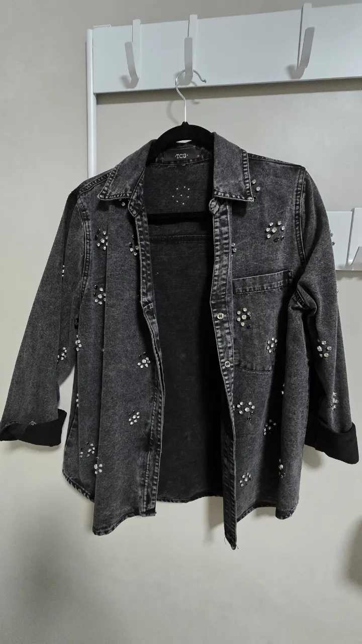 Campera de jean gris con detalles de strass y tachas en forma de flor. Posee cuello camisero, bolsillos en el frente y puños en las mangas. Ideal para un look canchero y con un toque de brillo.