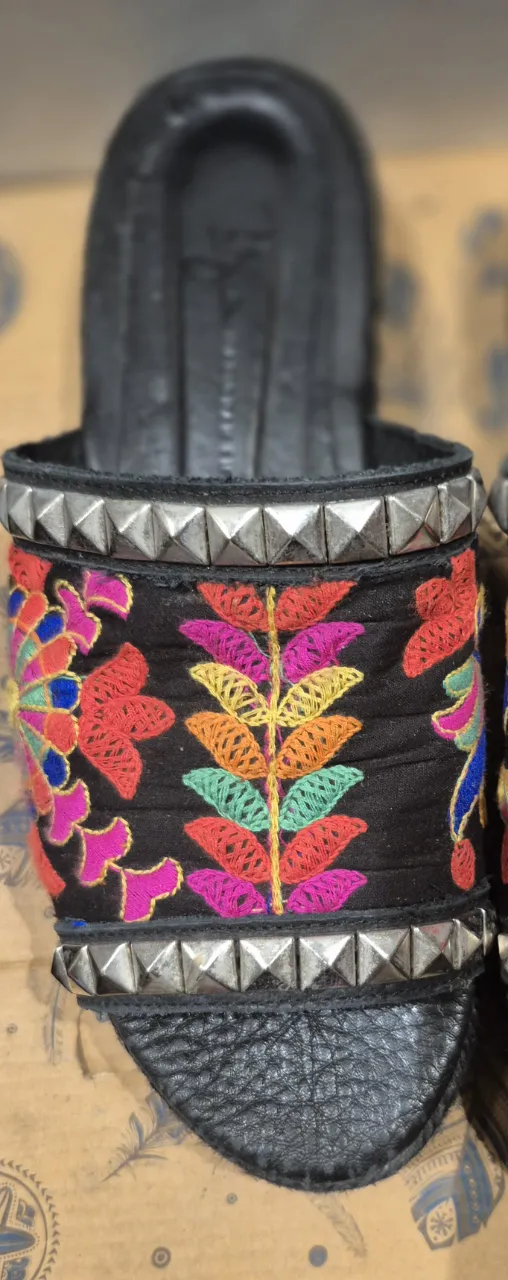 Sandalias de cuero negras con apliques bordados de flores y hojas en vivos colores. Tienen tachas plateadas alrededor de la base y la tira. Ideales para un look boho chic o rocker.