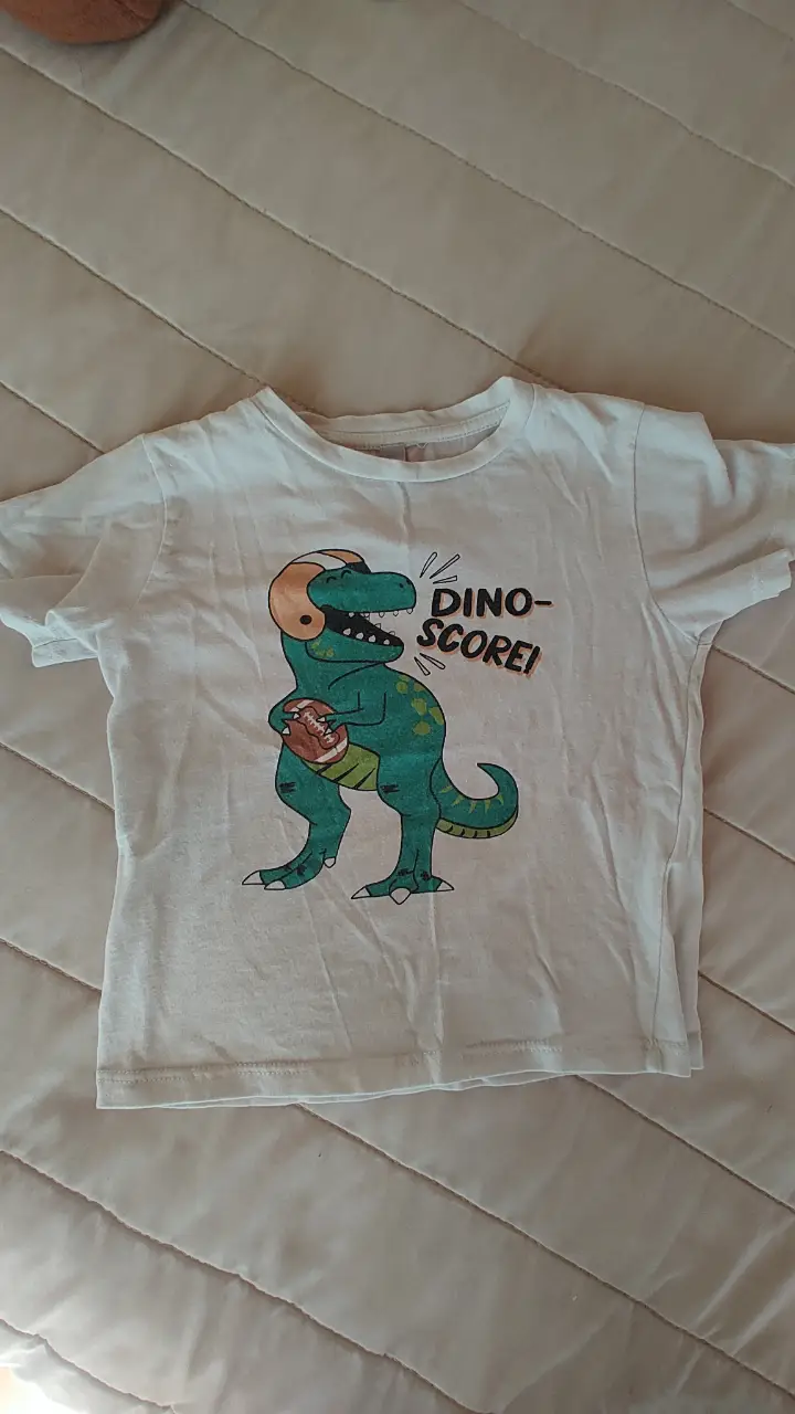 Remera blanca de niño con estampado de dinosaurio jugando al fútbol americano. Diseño divertido y cómodo para uso diario. Talle 3, tiene 34cm de largo y 29de ancho.