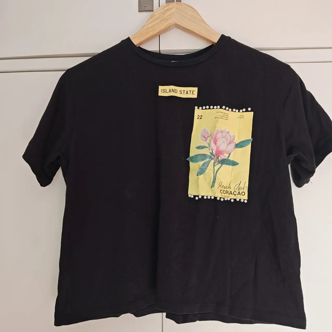 Remera negra de algodón con estampado floral en un bolsillo delantero. Diseño casual con detalles de perlas.