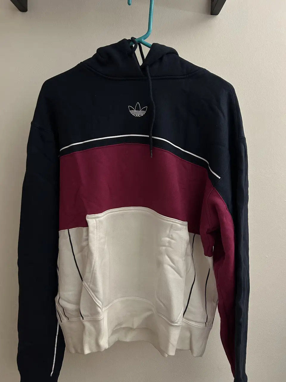 Buzo canguro Adidas de hombre, con diseño en bloques de color azul marino, borgoña y blanco. Posee el logo de la marca bordado en el pecho. Ideal para un look casual y deportivo.