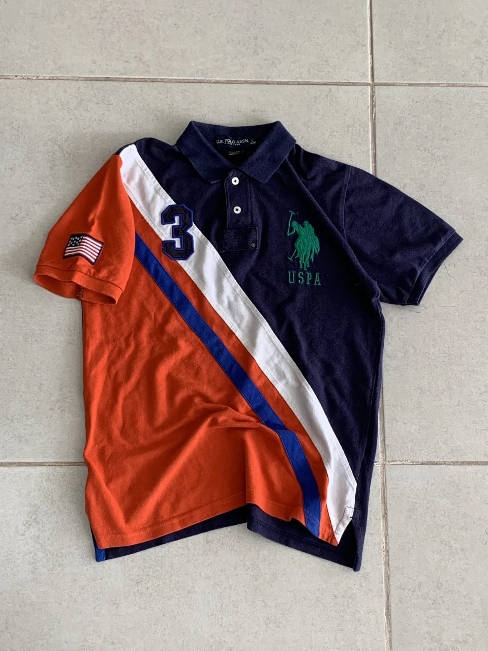 Remera tipo polo de manga corta, de color azul marino con detalles en naranja, blanco y azul. Posee un gran número 3 bordado en la parte frontal izquierda, y el logo de la marca bordado en verde en el lado derecho. En la manga izquierda tiene un parche con la bandera de Estados Unidos.