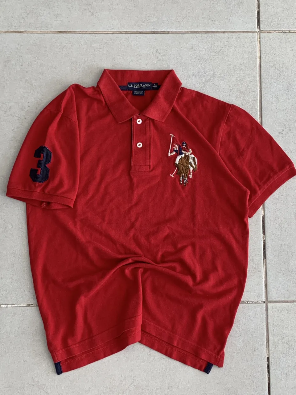 Remera polo roja con logo bordado de un jinete y caballo en el frente. Tiene un número 3 bordado en la manga izquierda. Cuello con botones.