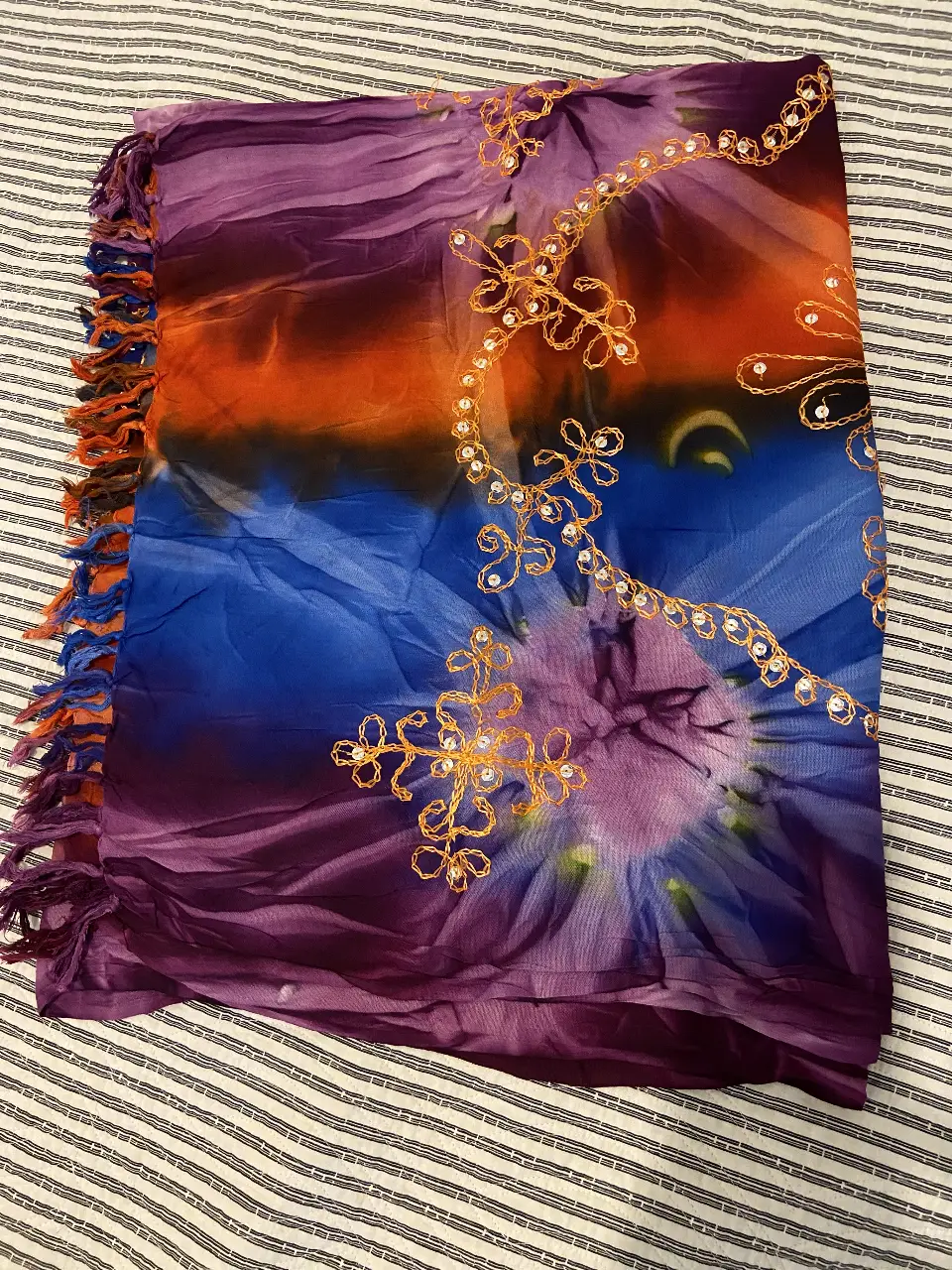 Pañuelo de seda con diseño tie-dye en tonos naranja, azul y violeta. Detalles bordados con brillos dorados. Ideal para complementar cualquier look.