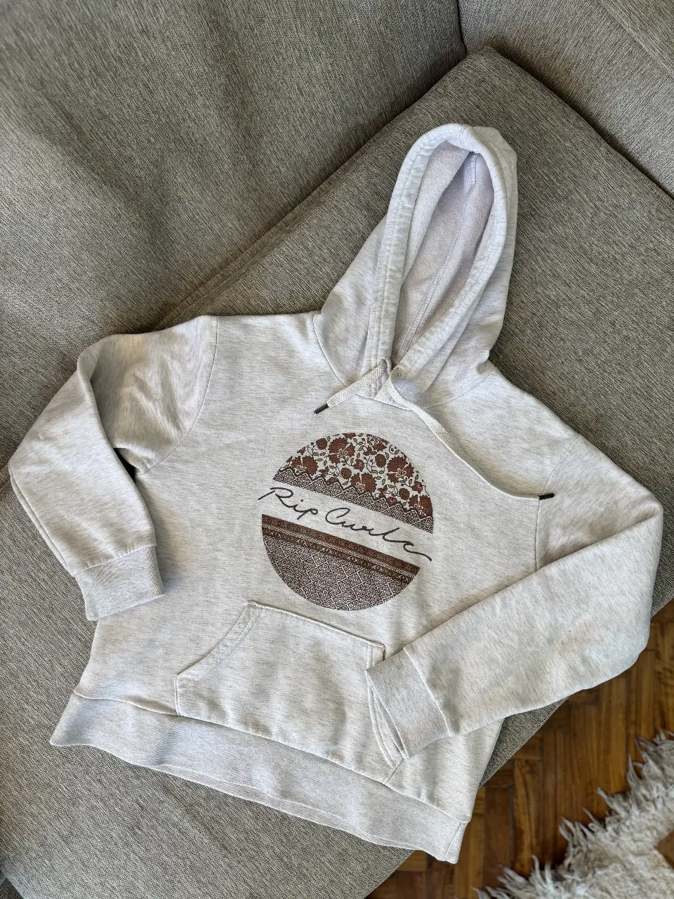 Buzo canguro Rip Curl gris con capucha y bolsillo delantero. Estampado circular en el pecho con detalles marrones y el logo de la marca. Ideal para un look casual y cómodo.