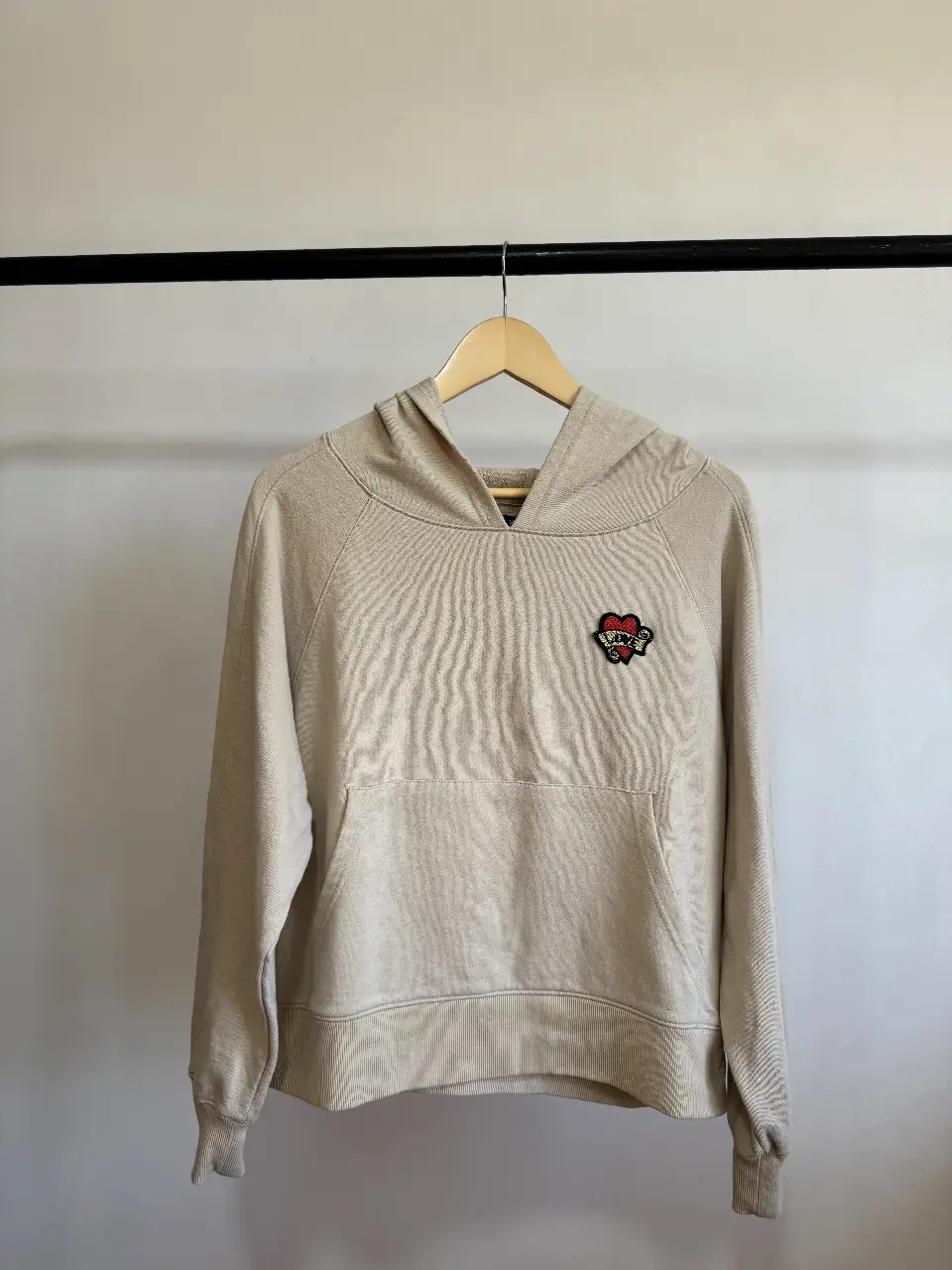 Buzo canguro de color crudo con capucha y bolsillo delantero. Tiene un parche con un corazón y detalles bordados. Ideal para un look casual y cómodo.