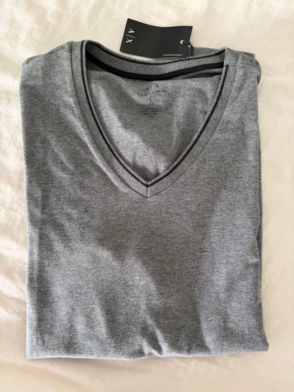 Remera gris de manga corta con cuello en V y detalles en negro en el cuello y mangas. Posee un logo de la marca en la parte superior de la espalda.