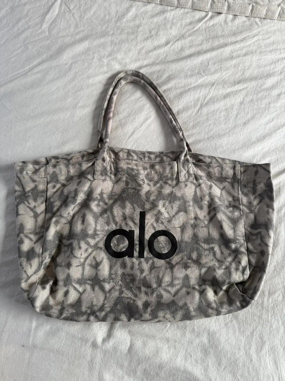 Bolso tote de la marca Alo, con estampado tie-dye en tonos grises y blancos. Ideal para llevar tus cosas con estilo y comodidad. lo use una sola vez y lo lave y se destiñó un poco la parte de colgar casi no se nota