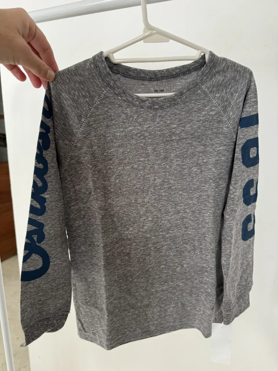 Buzo de cuello redondo para niño, color gris con mangas azules estampadas con la palabra "Oshkosh" en una manga y el número "6" en la otra. Ideal para un look casual y cómodo.