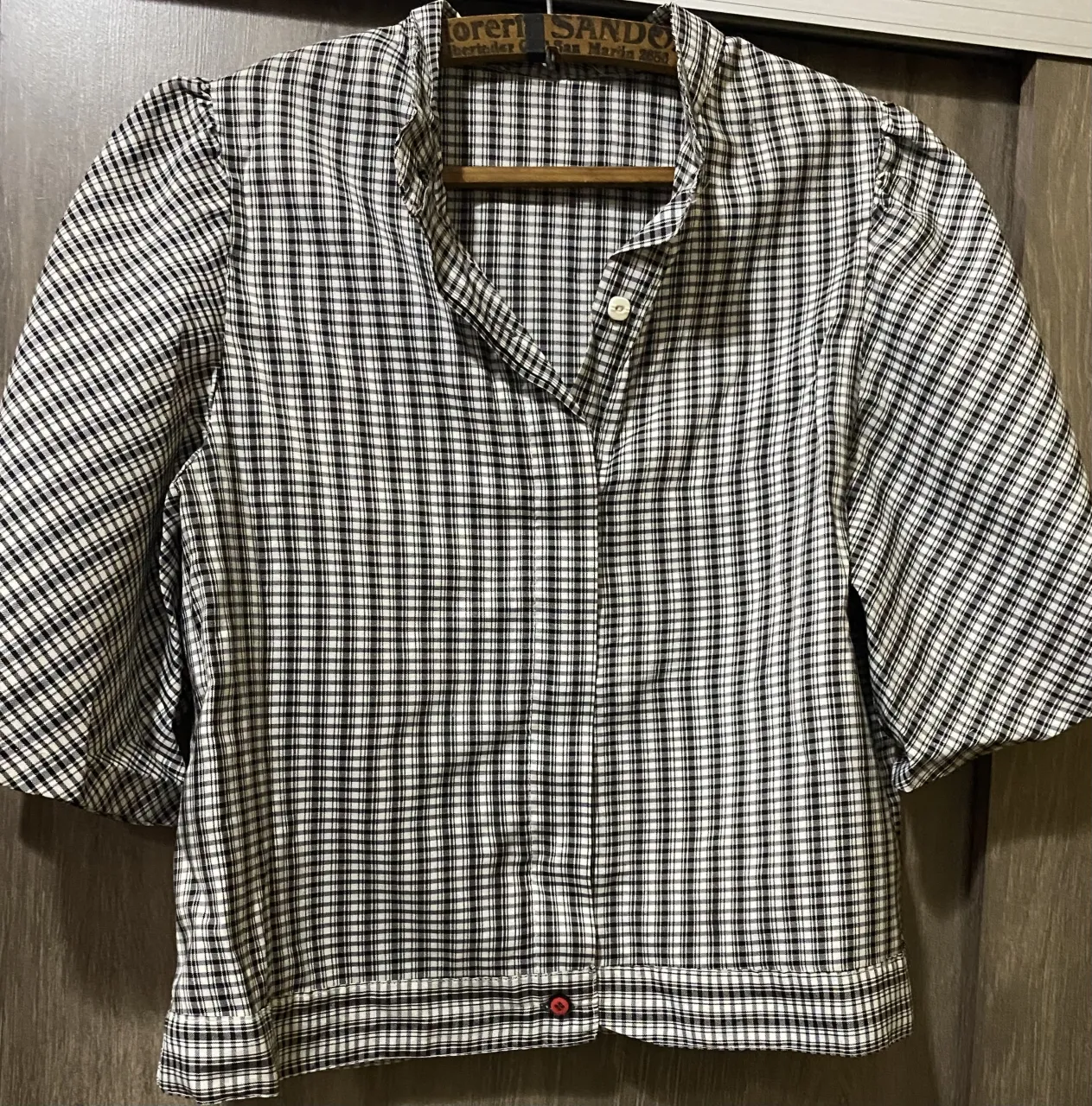 Remera a cuadros escoceses en blanco, negro y rojo. Posee cuello mao y botones al frente. Las mangas son cortas y con un detalle de cinta naranja en el lateral.