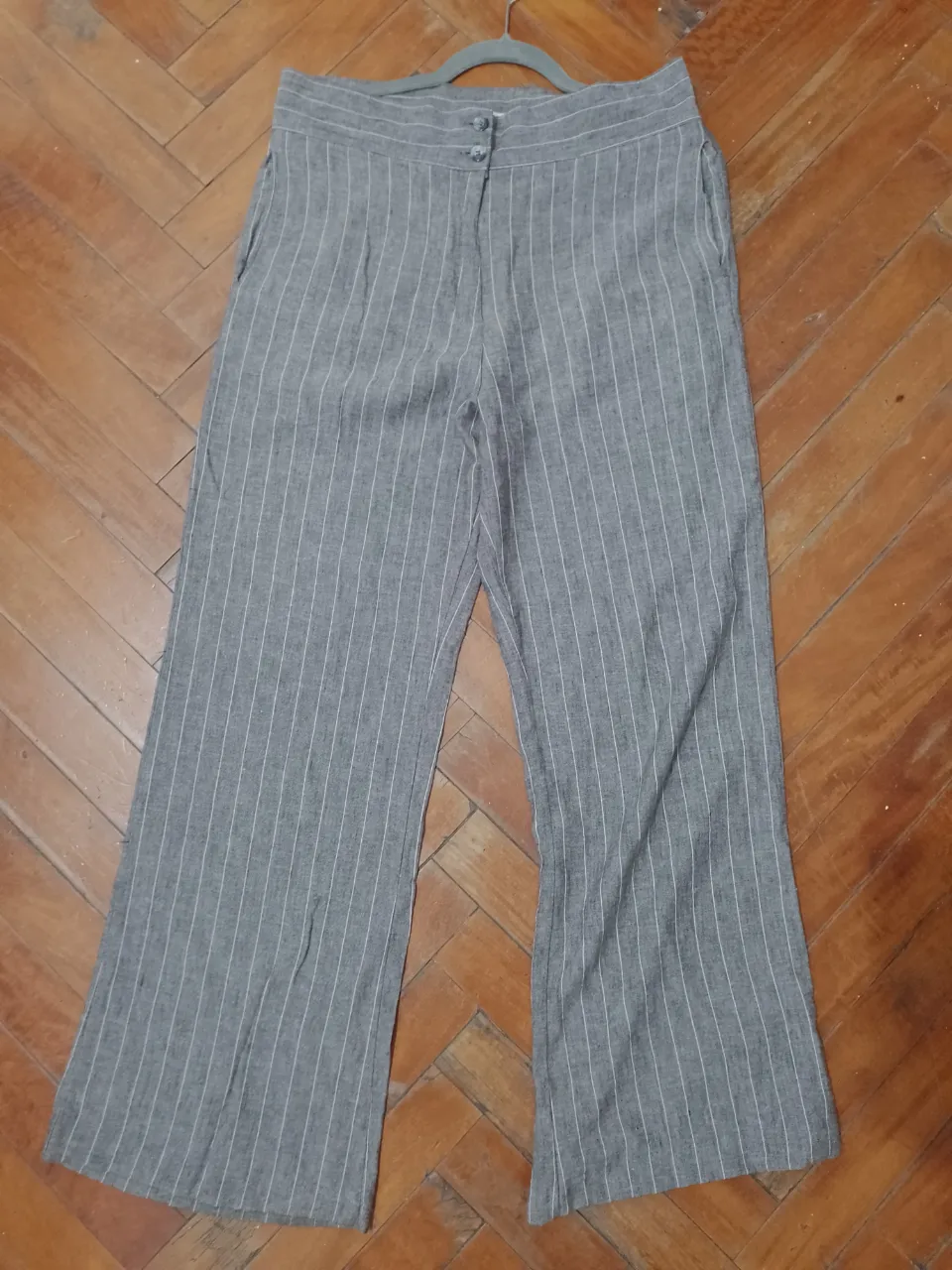 Pantalón sastrero de mujer, gris con rayas blancas verticales. Tiro alto, corte recto y ancho, con dos botones, cierre y bolsillos delanteros. Una única postura, está impecable. Ideal para un look elegante y cómodo.