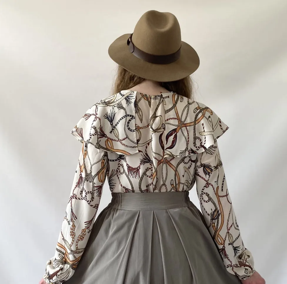 Blusa blanca con estampado de cadenas y hojas en tonos marrones, dorados y negros. Posee volados en el cuello y mangas largas con puños. Cierre en la espalda.