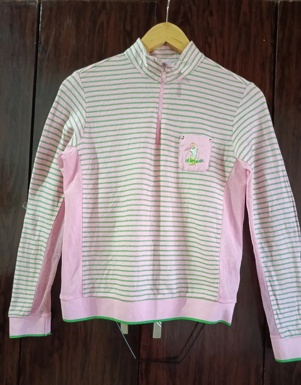 Remera deportiva rosa a rayas verdes con cuello mao y cierre. Tiene un bolsillo pequeño con un bordado de una figura femenina. Ideal para actividades al aire libre o un look casual.