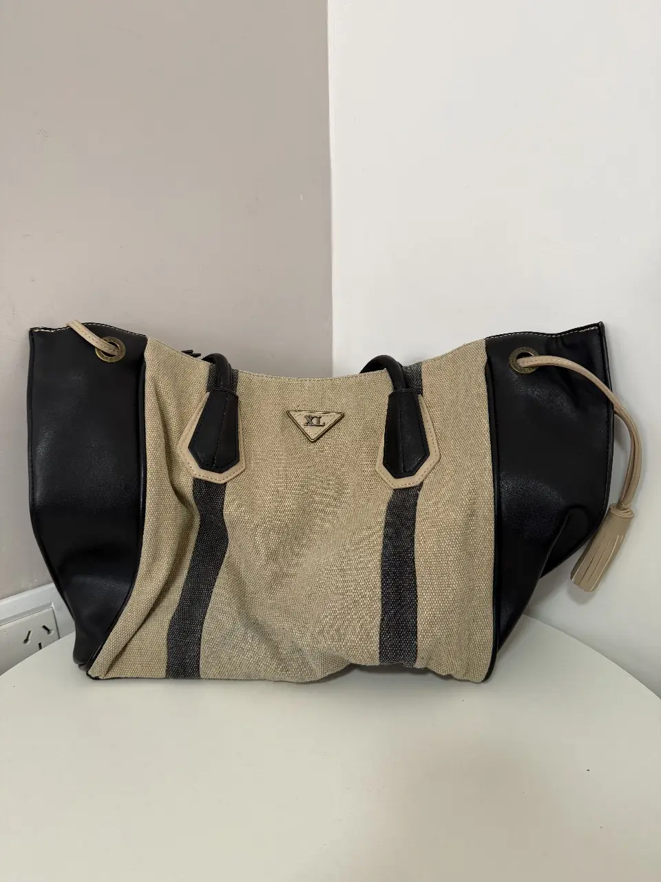 Bolso tote de mujer, con diseño bicolor en negro y beige. Confeccionado en lona y detalles en cuero sintético negro. Posee manijas superiores y un compartimento interno con cierre. Ideal para llevar tus pertenencias con estilo y comodidad.